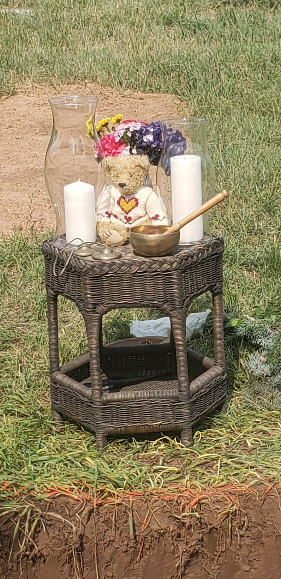 A woven altar table holding candles, a singing bowl and flowers beside a freshly dug grave